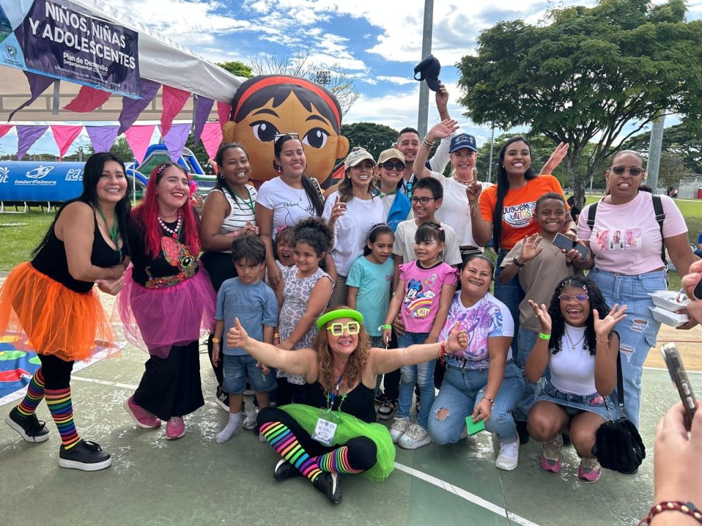 Fotografía grupal de una jornada recreativa en Palmira. Se observa a niñas, niños, familias y personal de apoyo posando con alegría en una cancha deportiva. Hay inflables, banderines coloridos y personas con atuendos festivos. Un cartel al fondo destaca el trabajo por la infancia y la adolescencia.