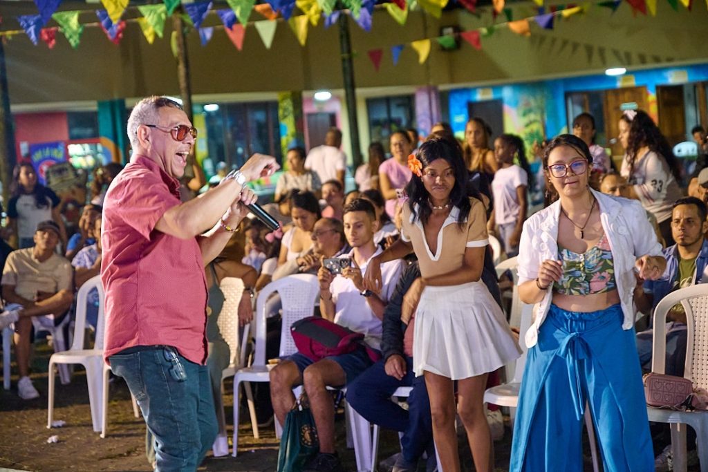 n hombre con camisa rosada y micrófono anima a una audiencia diversa durante un evento cultural nocturno bajo guirnaldas de colores. Dos jóvenes bailan con entusiasmo frente al público en un ambiente festivo.