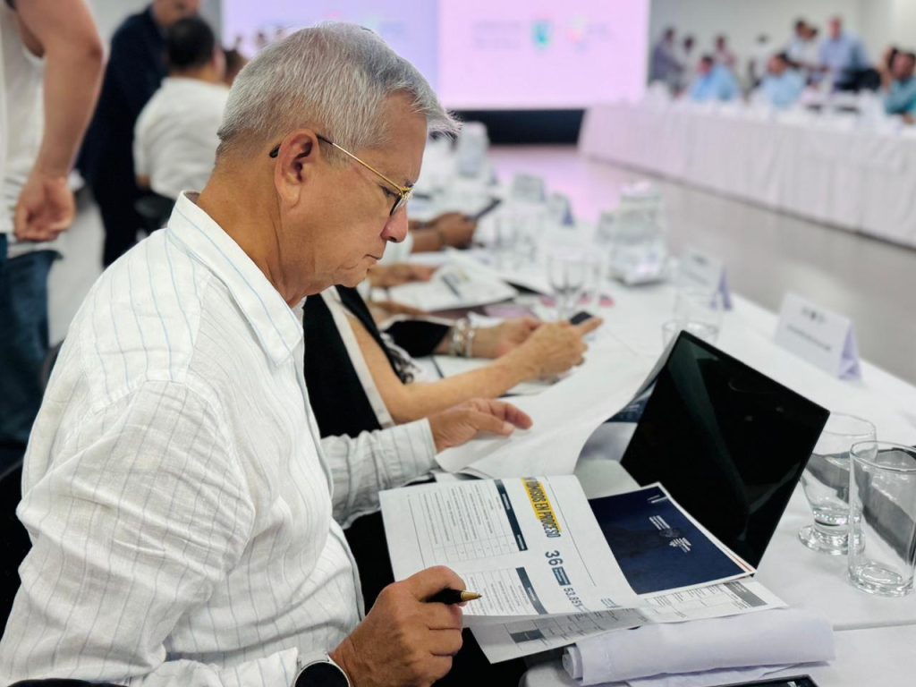 Primer plano de una persona mayor con gafas revisando documentos de indicadores y metas en una mesa de trabajo institucional. Al fondo, se observa un equipo de trabajo en una reunión de planificación estratégica