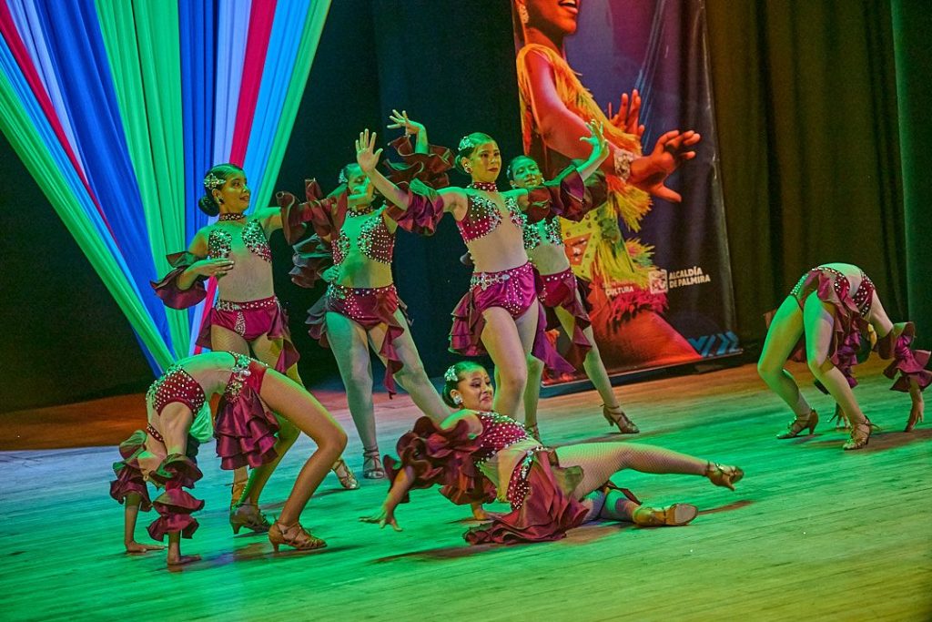Grupo de jóvenes bailarines con trajes color granate y lentejuelas realizando una coreografía dinámica en un escenario iluminado del Teatro Materon