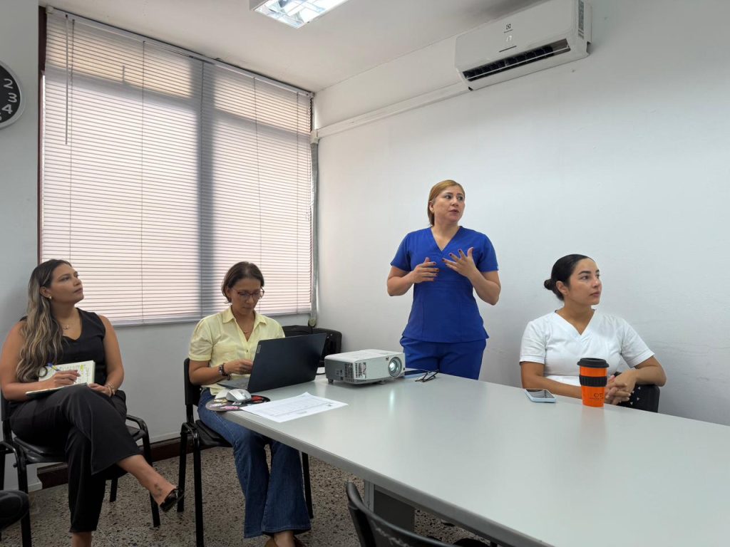 Cuatro profesionales en una reunión de trabajo; una persona con uniforme médico expone frente a sus colegas en una oficina con proyector
