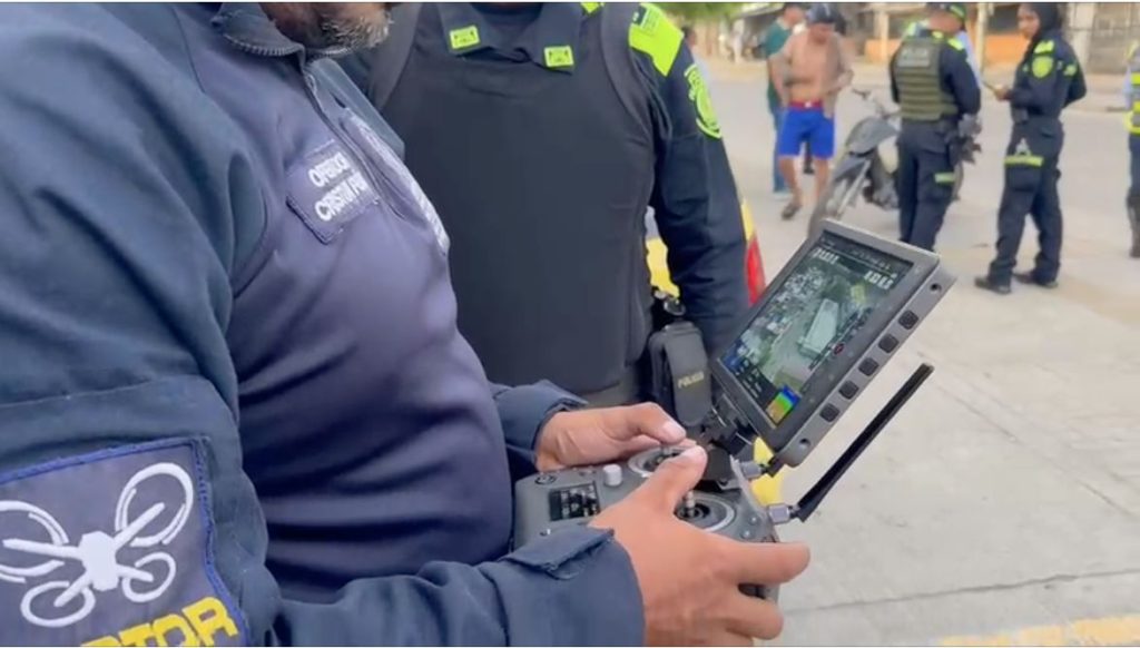 Primer plano de una persona de la policía operando un control remoto con pantalla incorporada para pilotar un dron. En el brazo de su chaqueta se observa un parche con el ícono de un dron. Al fondo, otras personas uniformadas y civiles se encuentran en una calle.