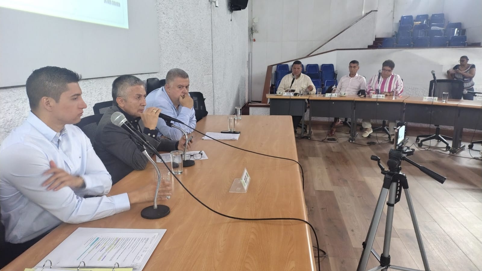 Un grupo de personas participa en una mesa de diálogo legislativa en un salón institucional. En primer plano, tres hombres están sentados frente a micrófonos en una mesa larga de madera; al fondo, otras personas ocupan una mesa secundaria. Un trípode con un dispositivo móvil registra el evento.