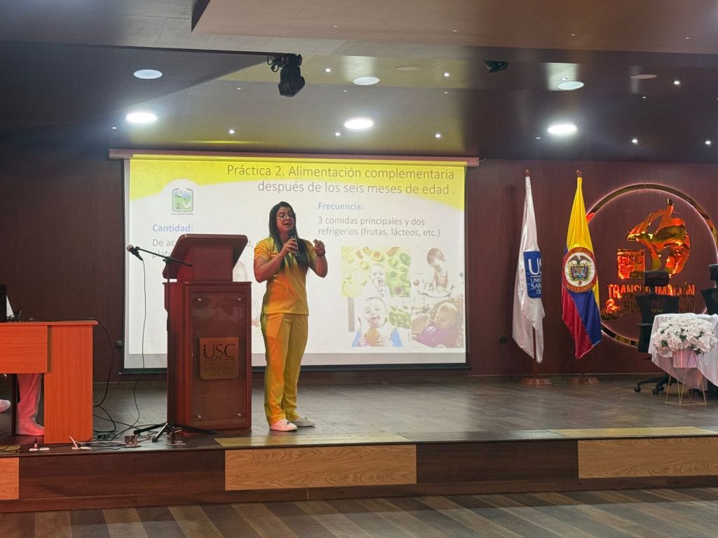 Una persona conferencista con uniforme amarillo expone sobre alimentación complementaria para bebés en un auditorio de la Universidad Santiago de Cali (USC). A su lado hay un podio de madera con el logo de la universidad y una pantalla de proyección con información técnica.