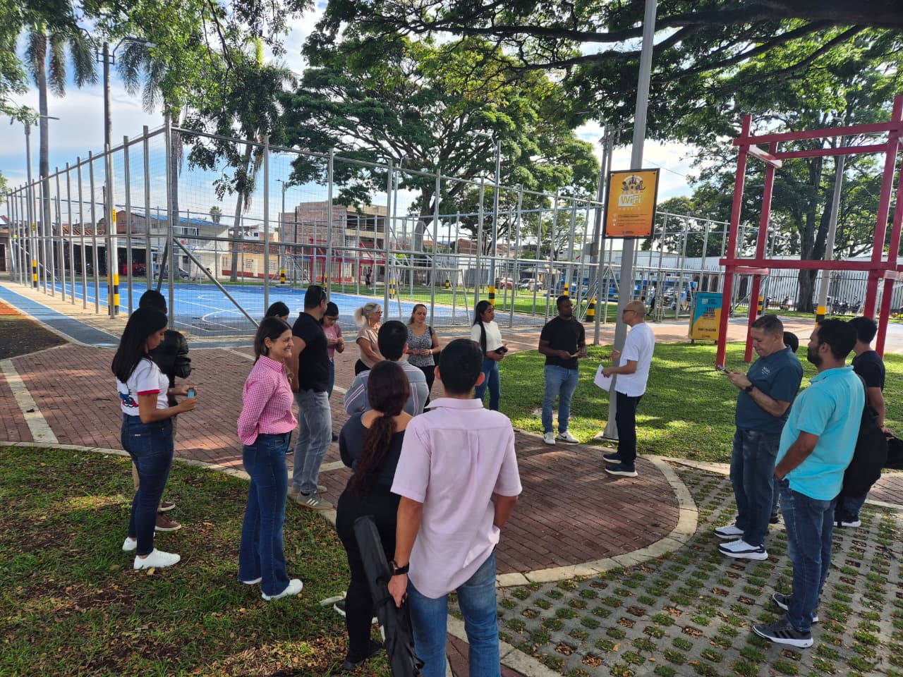 Un grupo de personas diversas se reúne en un parque frente a una cancha deportiva azul. Al fondo se ven árboles grandes y estructuras metálicas, mientras el grupo escucha con atención a una persona que sostiene unos documentos.