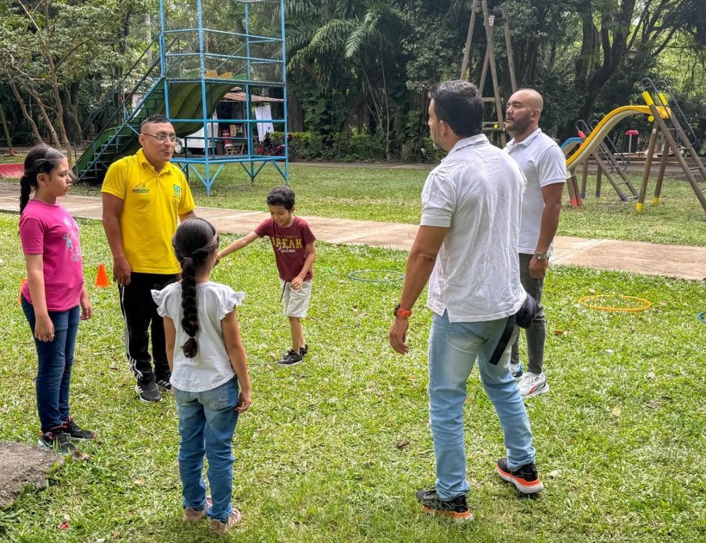 Un grupo de personas, incluyendo infantes y personas adultas, participan en una actividad recreativa al aire libre en un parque con zonas verdes y juegos infantiles