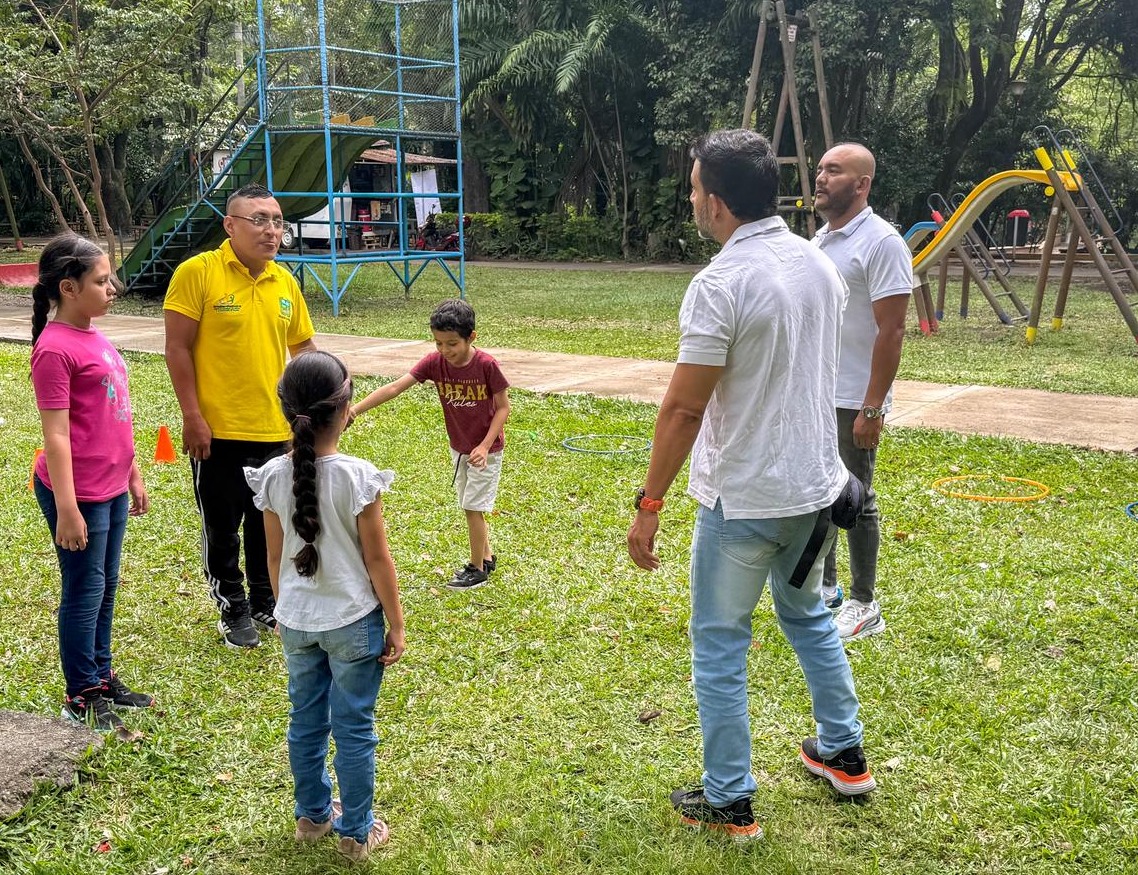 Un grupo de personas, incluyendo infantes y personas adultas, participan en una actividad recreativa al aire libre en un parque con zonas verdes y juegos infantiles