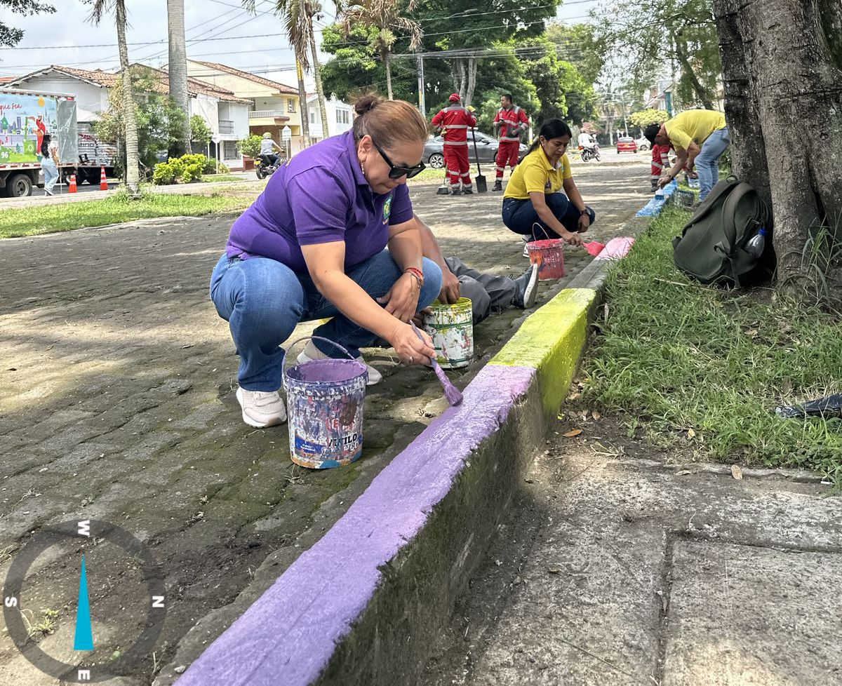 Una persona con camiseta morada y gafas de sol pinta el bordillo de una calle de color lavanda. Al fondo, otras personas colaboran en la tarea pintando tramos de color amarillo y rosa en una zona arbolada.