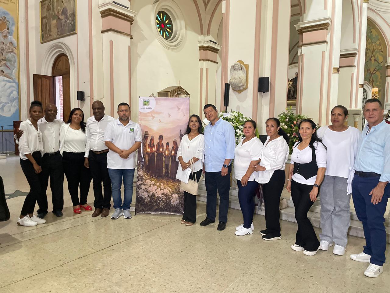 Un grupo de doce personas posa junto a un pendón informativo en el interior de una iglesia. La mayoría viste prendas blancas y oscuras, y se encuentran situadas frente a un mural que muestra a personas sosteniendo velas