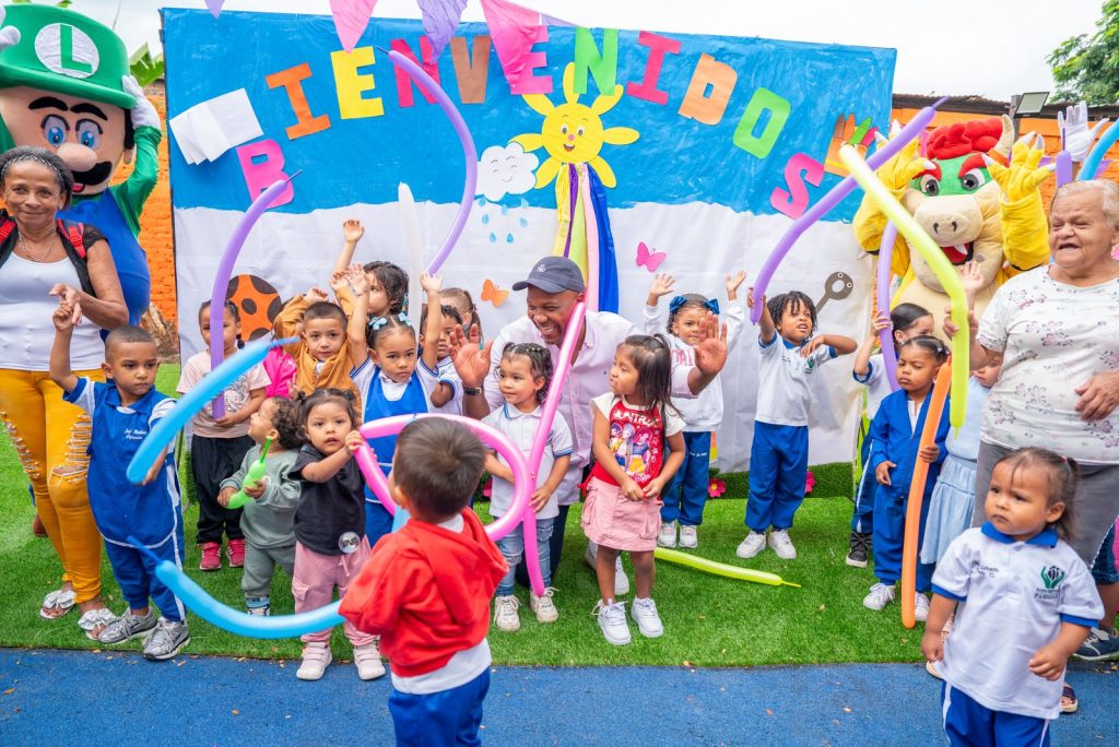 Grupo de niños y adultos celebrando con globos frente a un mural de "Bienvenidos" junto al alcalde de Palmira, Víctor Ramos
