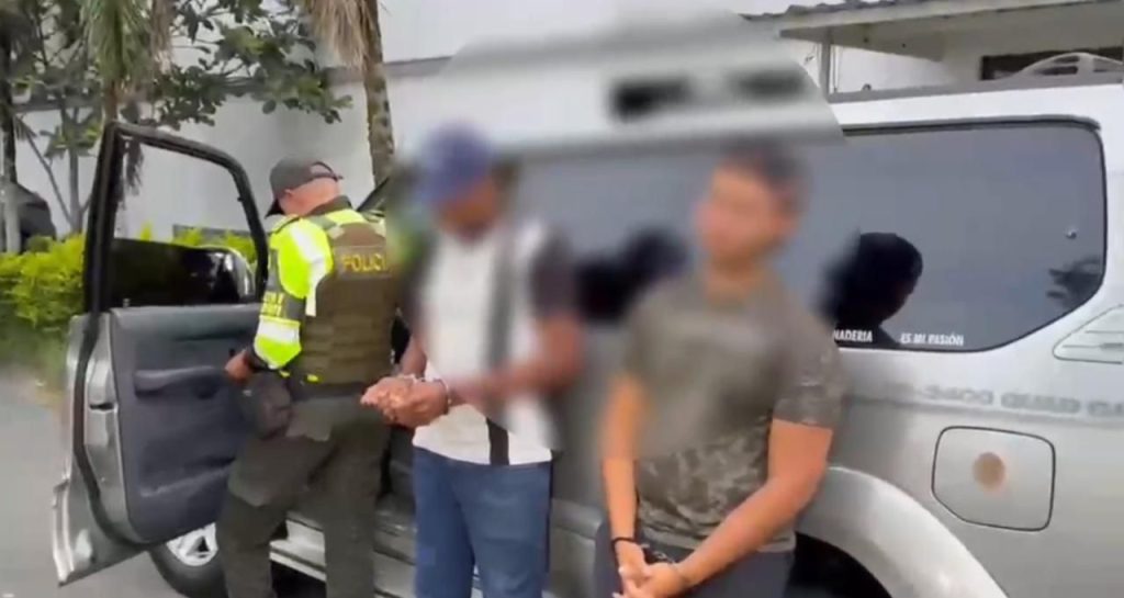 Personal policial realizando un procedimiento junto a dos personas bajo custodia oficial al lado de un vehículo gris. Las identidades de las personas civiles están protegidas mediante difuminado