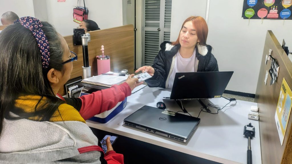 Una mujer entrega su documento de identidad a una funcionaria que la atiende frente a un computador en una oficina