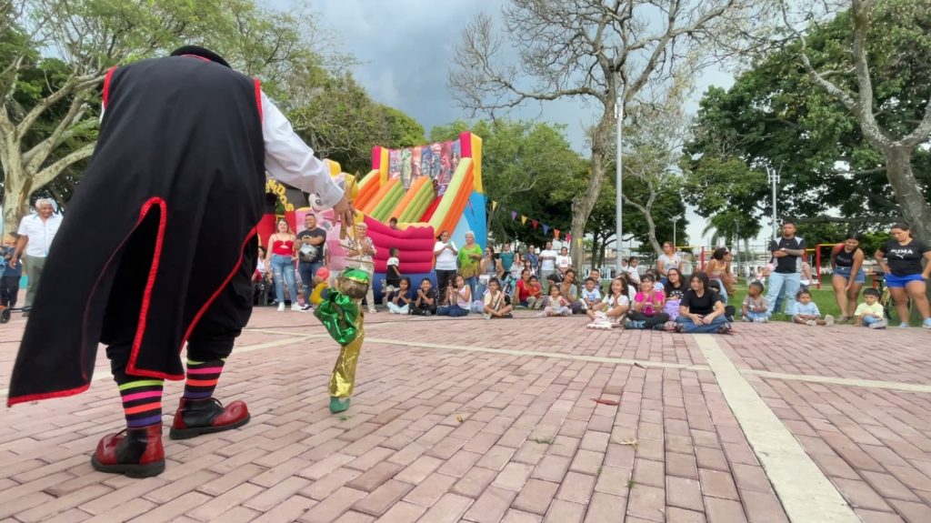 Persona vestida de payaso realizando un espectáculo de marionetas frente a un público infantil y adulto sentado en centro cultural y deportivo El Prado. De fondo se observa un inflable colorido y árboles