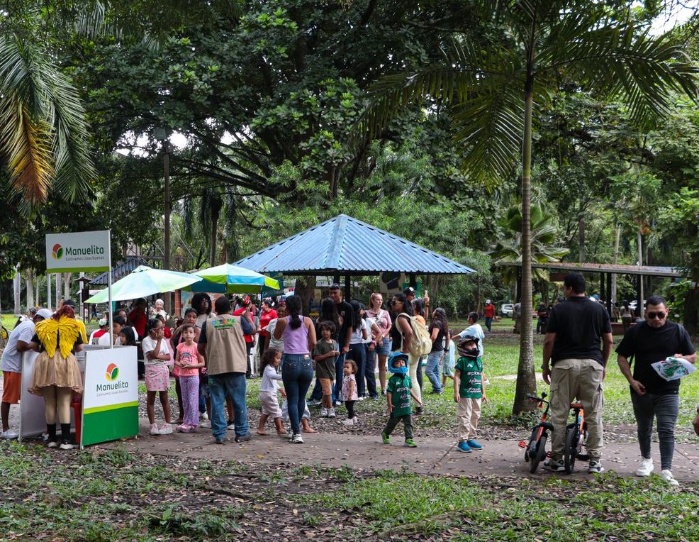Grupo diverso de personas, incluyendo familias, niñas y niños, participando en un evento comunitario al aire libre en el Bosque Municipal con stands de Manuelita y un quiosco central