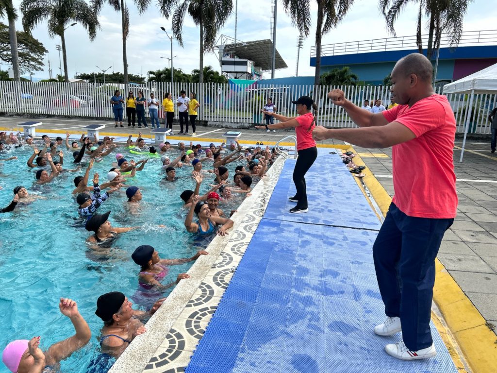 Un grupo numeroso de personas mayores realiza ejercicios de gimnasia acuática en una piscina pública, guiados por dos instructores desde el borde