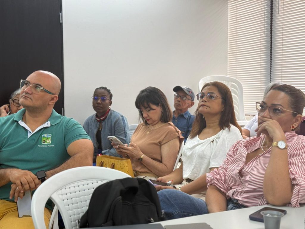 Grupo de personas, hombres y mujeres de diversas edades, sentados en una reunión o conferencia. En primer plano, una persona con polo verde escucha atentamente, mientras otras toman notas o usan sus teléfonos móviles