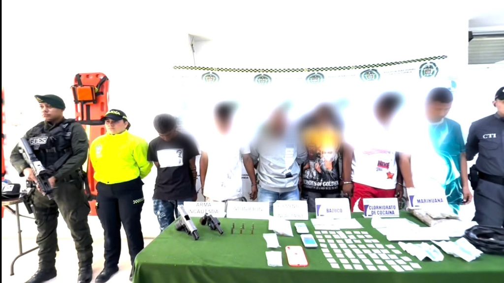 Siete personas con rostros difuminados permanecen de pie tras una mesa con elementos incautados, custodiadas por tres integrantes de las fuerzas de seguridad en Colombia. Sobre la mesa se observan armas, sustancias y otros indicios