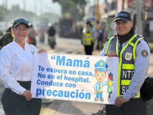 Dos agentes de tránsito, una mujer y un hombre, sostienen un cartel de seguridad vial dirigido a las madres. El mensaje dice: 'Mamá te espera en casa, no en el hospital. Conduce con precaución'
