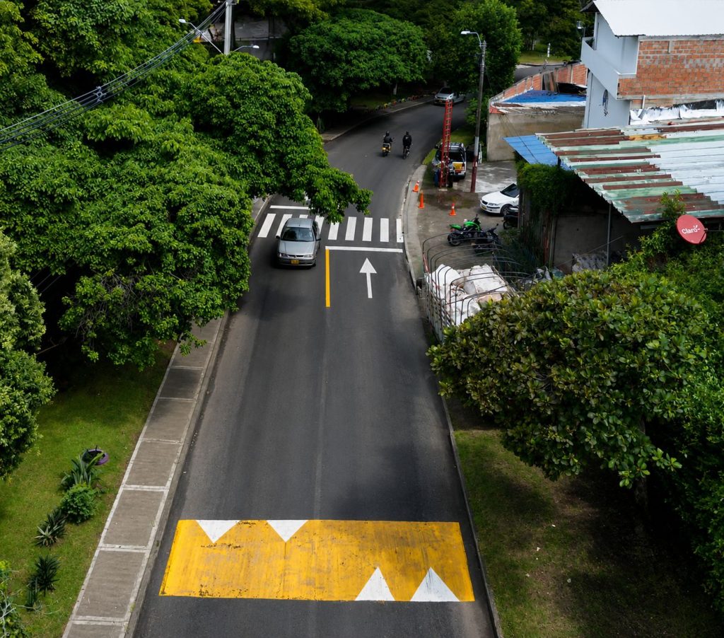 Vista aérea de una calle asfaltada con señalización vial renovada. Se observa un resalto amarillo con triángulos blancos en primer plano, seguido de una flecha de dirección, una línea de pare y un paso peatonal tipo cebra