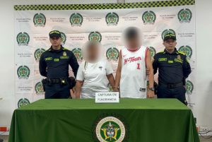 Dos personas con rostros difuminados, un hombre y una mujer, permanecen de pie tras una mesa con un cartel que dice 'Captura en flagrancia'. Están custodiadas por dos agentes de la Policía Nacional de Colombia frente a un fondo institucional