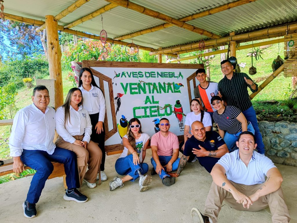 Un grupo diverso de once personas posa sonriente frente a un gran letrero que dice 'Aves de Niebla - Ventanal al Cielo'. Se encuentran en una estructura abierta de madera y guadua en un entorno rural.