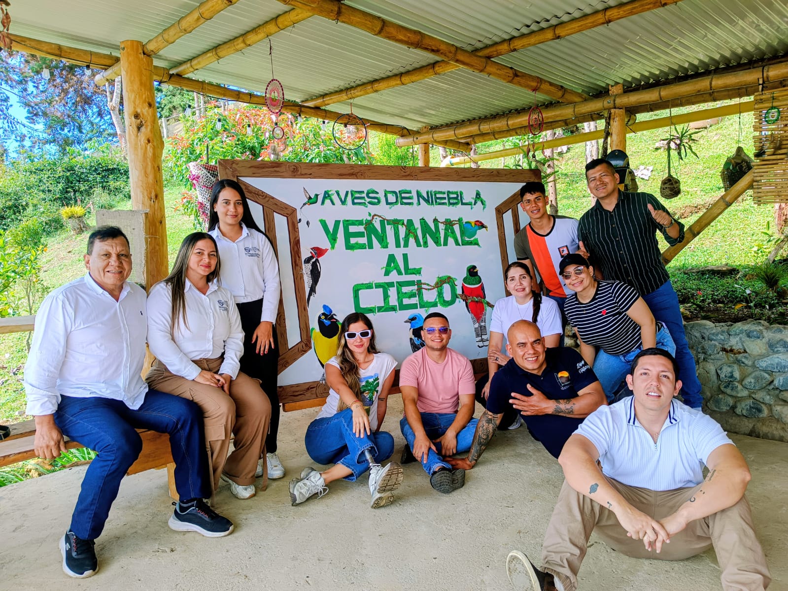 Un grupo diverso de once personas posa sonriente frente a un gran letrero que dice 'Aves de Niebla - Ventanal al Cielo'. Se encuentran en una estructura abierta de madera y guadua en un entorno rural.