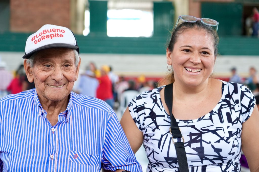 Un hombre mayor y una mujer joven sonríen a la cámara en un evento comunitario. El hombre lleva una gorra blanca con la etiqueta '#LoBuenoSeRepite' y una camisa de rayas azules, mientras la mujer viste una blusa con estampado geométrico