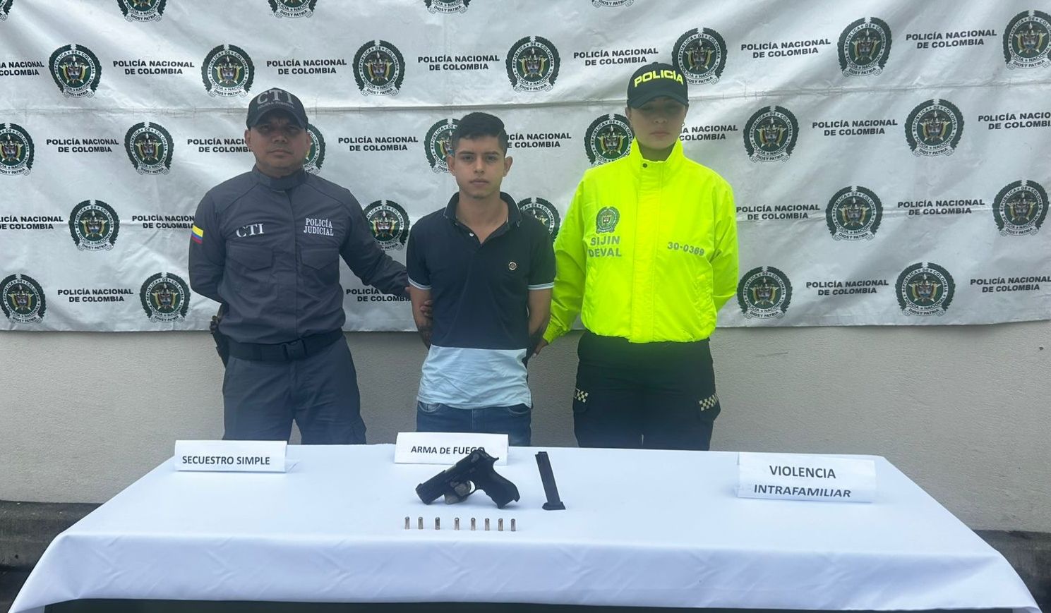 Fotografía institucional de la Policía Nacional de Colombia que muestra a una persona bajo custodia, flanqueada por una mujer policía y un agente del CTI. Sobre una mesa frente a ellos se exhiben una pistola, un cargador y cartuchos, junto a letreros que indican los delitos de secuestro simple, violencia intrafamiliar y porte de arma de fuego