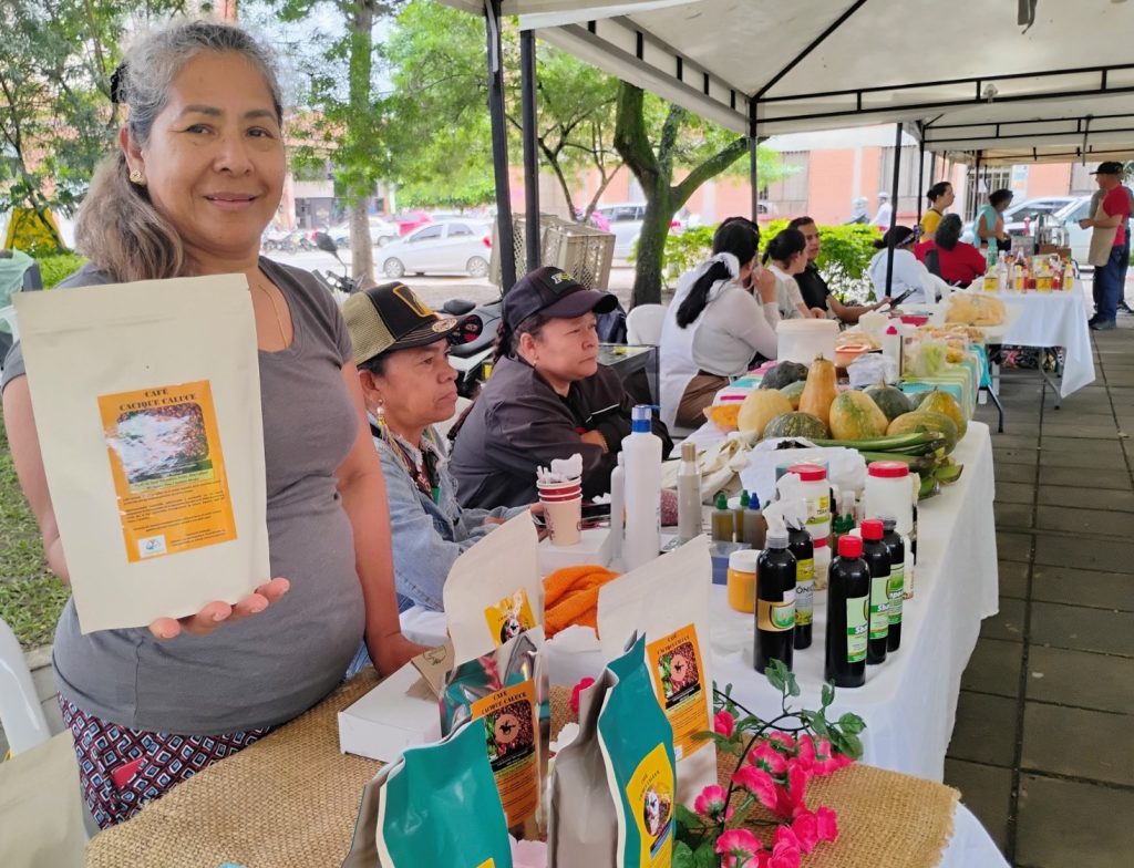 Una mujer sonriente en un mercado al aire libre sostiene un paquete de café artesanal frente a su puesto. Al fondo, otras personas atienden mesas con productos agrícolas y artesanías bajo carpas blancas.