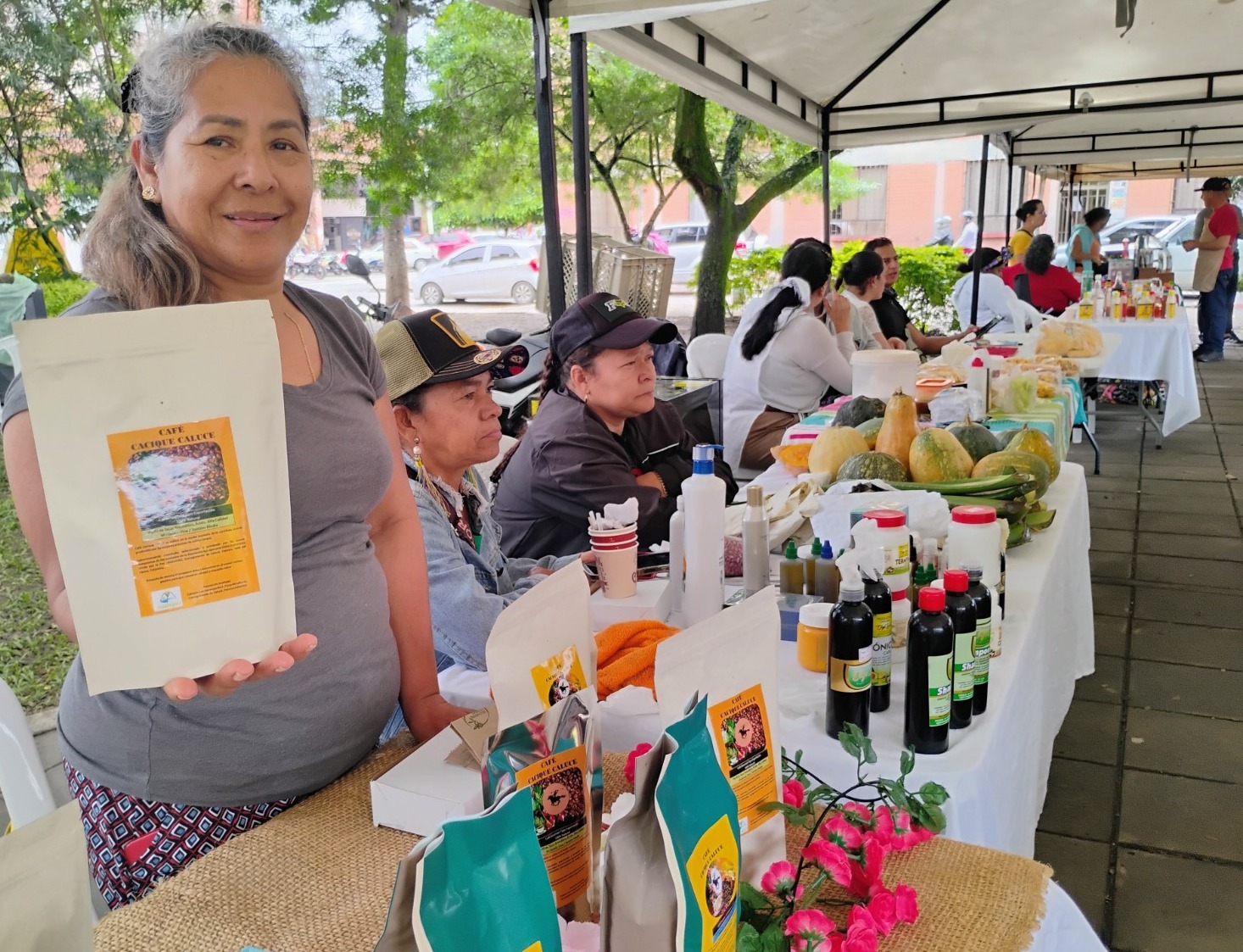 Una mujer sonriente en un mercado al aire libre sostiene un paquete de café artesanal frente a su puesto. Al fondo, otras personas atienden mesas con productos agrícolas y artesanías bajo carpas blancas.