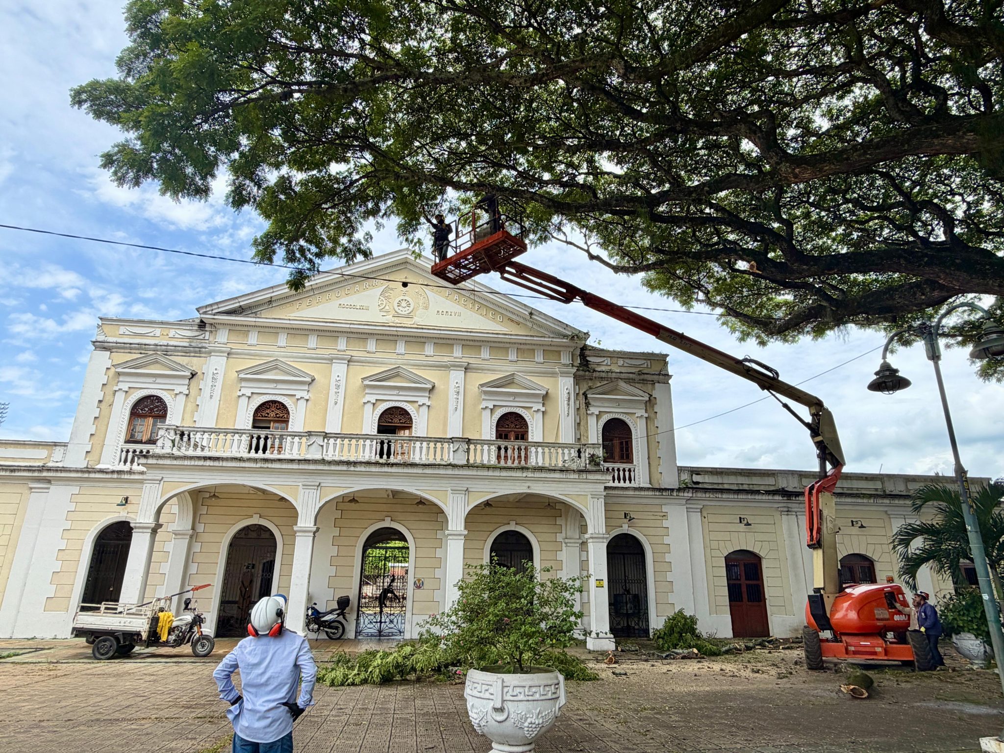 Personal técnico realiza la poda de un gran árbol utilizando una plataforma elevadora frente a la fachada de un edificio histórico de la Antigua Estación del Ferrocarril