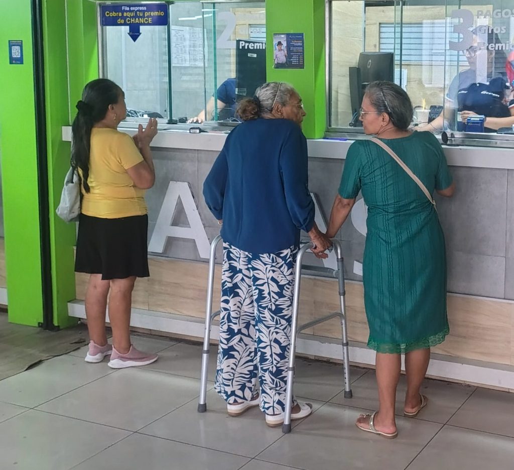 Tres personas adultas esperan frente a las ventanillas de un centro de atención o pagos. Una de ellas, con cabello cano, utiliza un andador metálico para apoyarse mientras conversa con otra persona a su lado