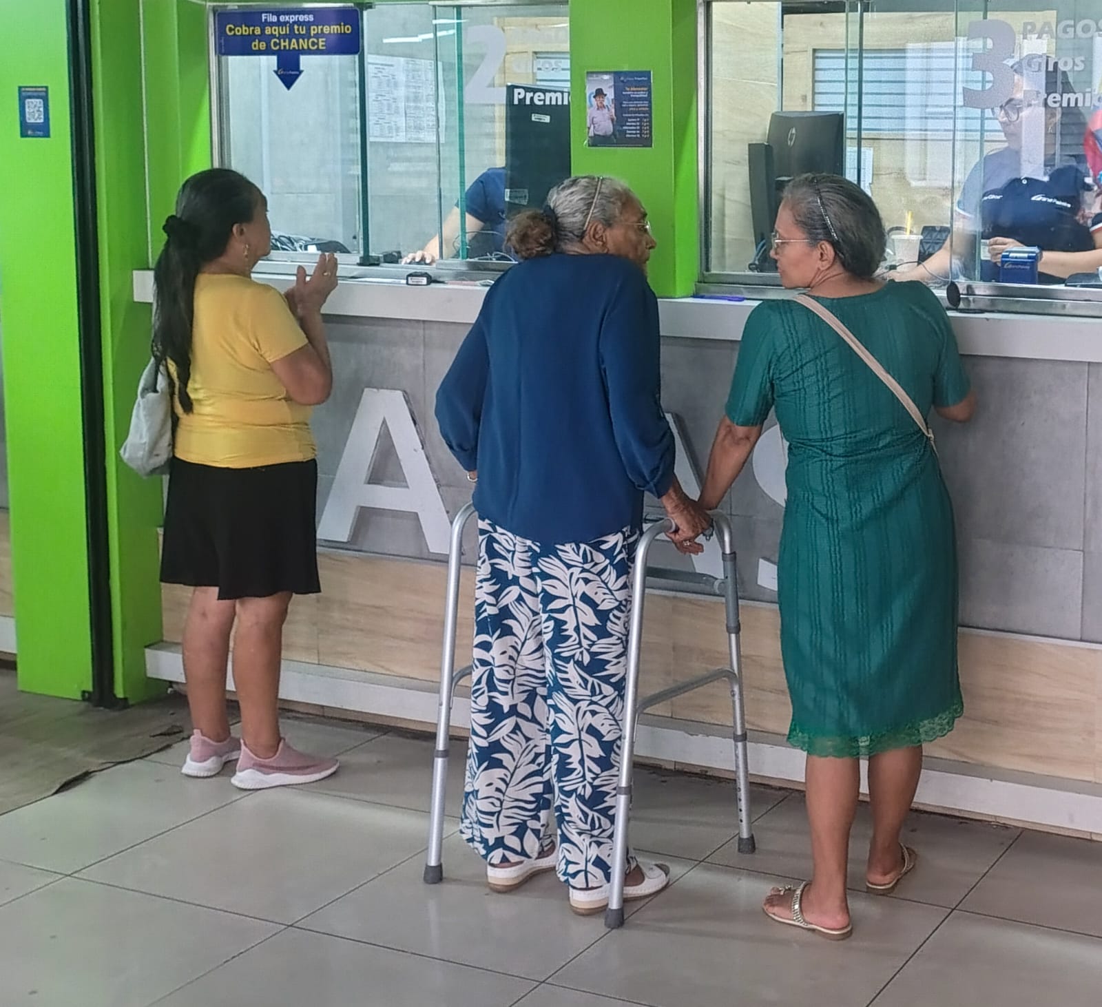Tres personas adultas esperan frente a las ventanillas de un centro de atención o pagos. Una de ellas, con cabello cano, utiliza un andador metálico para apoyarse mientras conversa con otra persona a su lado