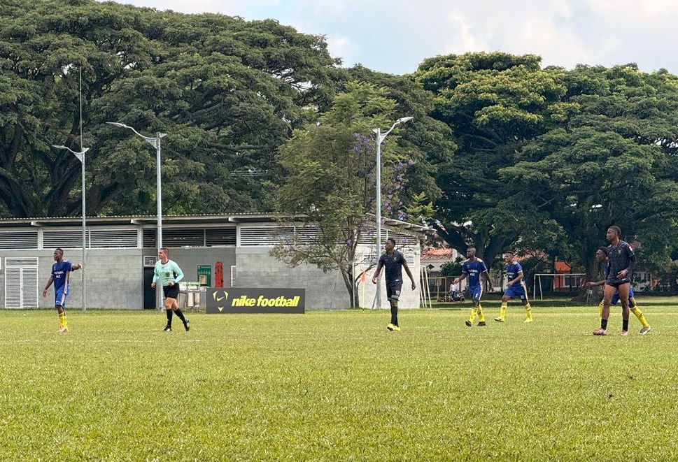 otografía de un partido de fútbol amateur en una cancha de césped natural, rodeada de árboles frondosos y postes de luz. En el campo se observan varias personas jugando con uniformes oscuros y azules, junto a un integrante del cuerpo arbitral con camiseta clara, en un ambiente deportivo al aire libre