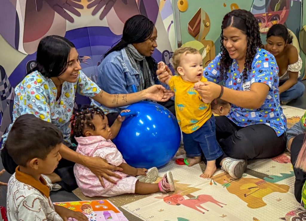 Fotografía en plano medio de una sesión de estimulación temprana en interiores. Profesionales de la salud o educación interactúan con alegría junto a infantes y sus familias sobre un tapete infantil con ilustraciones. En el centro, un bebé en camiseta amarilla da pasos con apoyo, mientras otra infancia se sienta junto a un gran balón terapéutico azul.