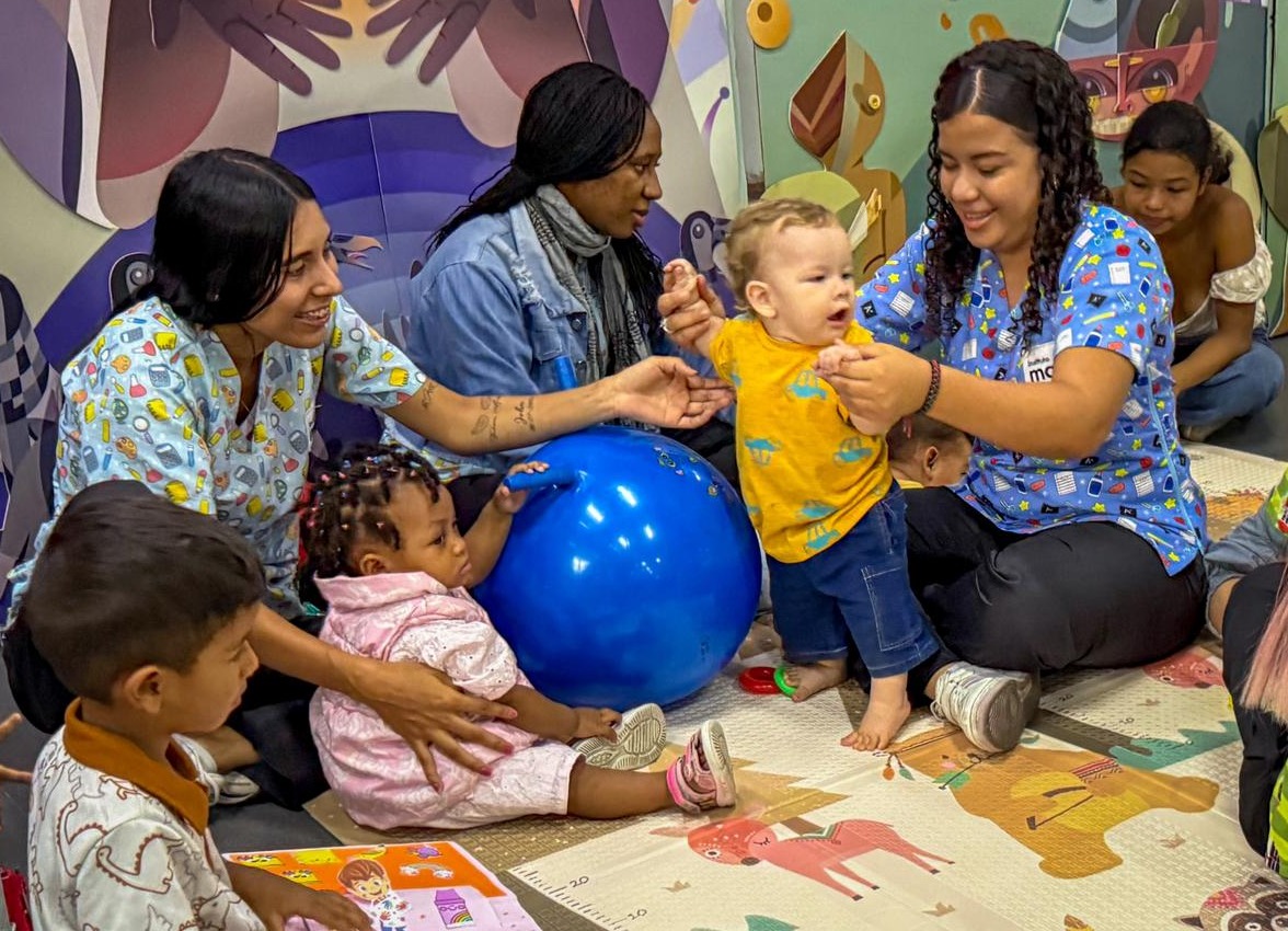 Fotografía en plano medio de una sesión de estimulación temprana en interiores. Profesionales de la salud o educación interactúan con alegría junto a infantes y sus familias sobre un tapete infantil con ilustraciones. En el centro, un bebé en camiseta amarilla da pasos con apoyo, mientras otra infancia se sienta junto a un gran balón terapéutico azul.