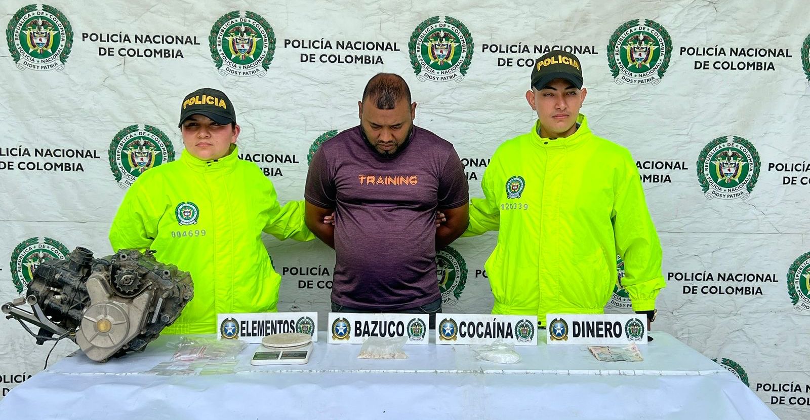 Fotografía oficial de la Policía Nacional de Colombia donde dos agentes escoltan a un hombre bajo custodia frente a una mesa con elementos incautados: un motor de motocicleta, bolsas con sustancias etiquetadas como bazuco y cocaína, dinero en efectivo y una báscula gramera