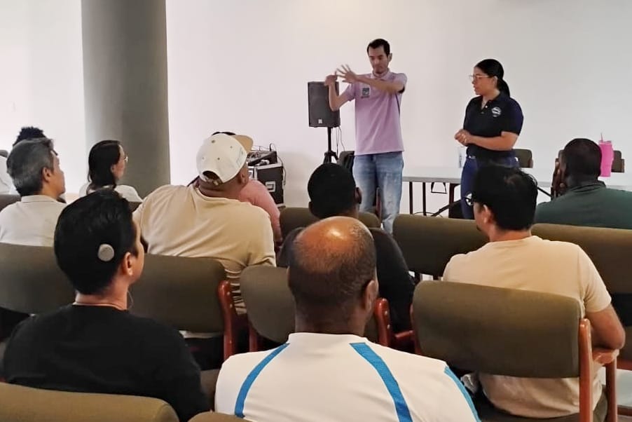 Una mujer expone y un interprete de señas realiza traducción de pie frente a un grupo de personas sentadas en un auditorio. El hombre realiza señas con las manos mientras la mujer observa
