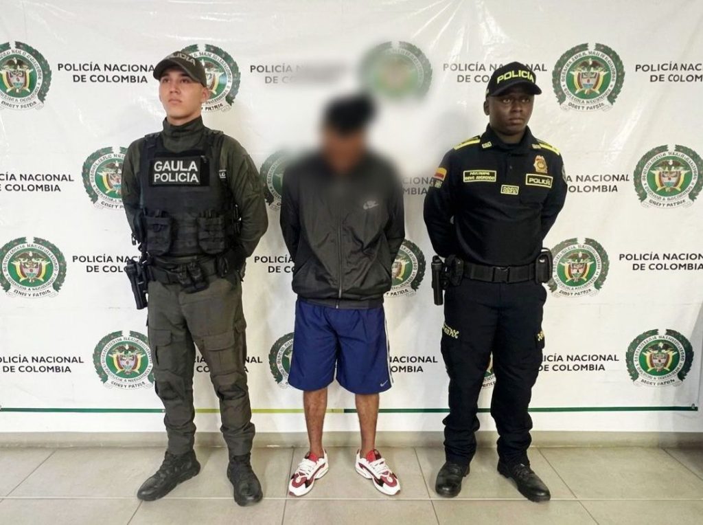 Un hombre de pie con el rostro difuminado permanece entre dos agentes de la Policía Nacional de Colombia frente a un muro con logotipos institucionales.