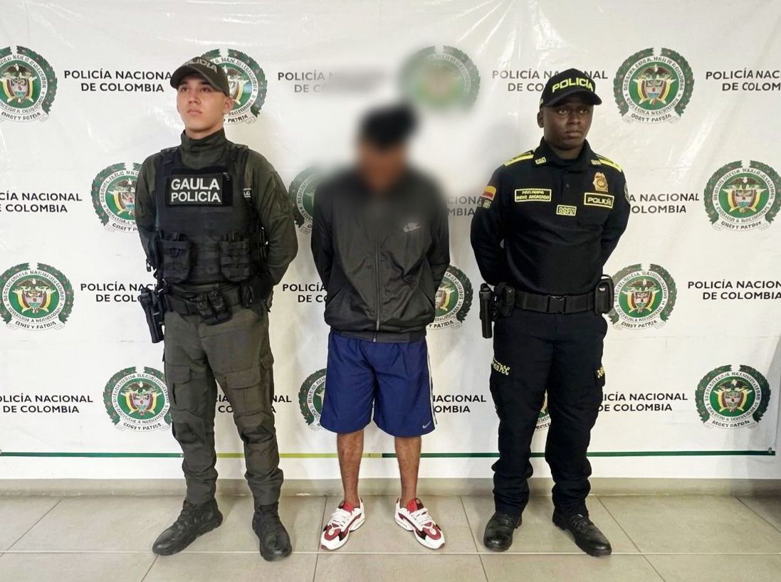 Un hombre de pie con el rostro difuminado permanece entre dos agentes de la Policía Nacional de Colombia frente a un muro con logotipos institucionales.