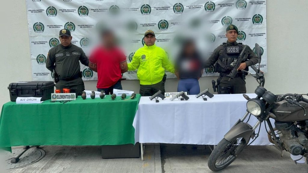 Fotografía institucional de la Policía Nacional de Colombia que muestra la presentación pública de dos personas bajo custodia, acompañadas por agentes de policía frente a un muro con logotipos oficiales, junto a elementos incautados sobre mesas y una motocicleta