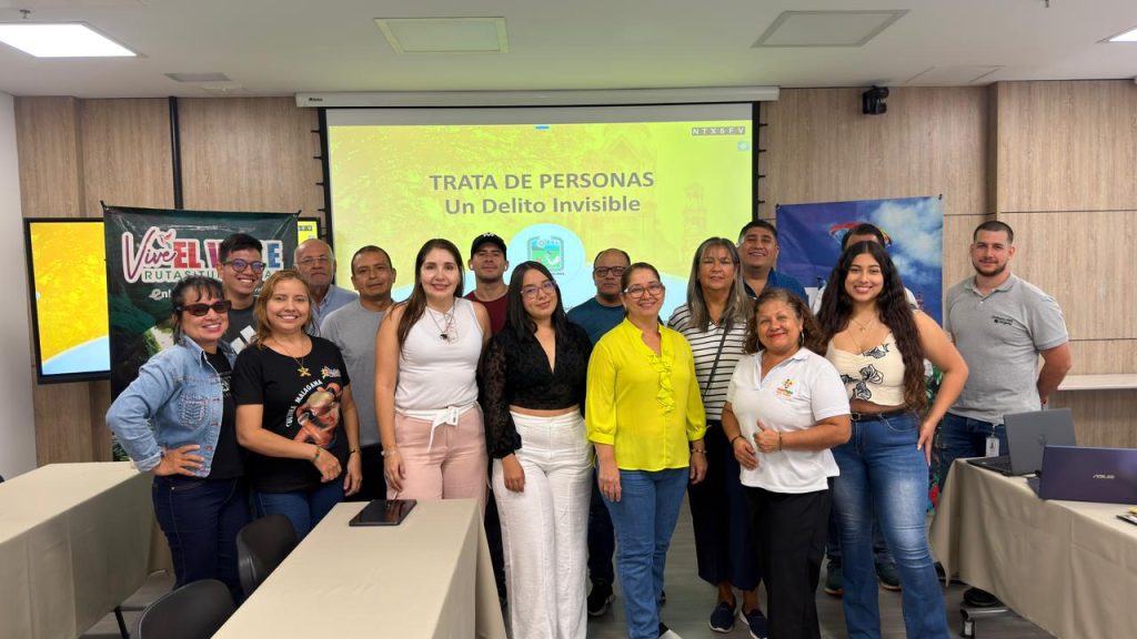 Fotografía de un grupo de trece personas de diversos géneros y edades posando juntas en una sala de capacitación frente a una pantalla de proyección que dice "TRATA DE PERSONAS Un Delito Invisible"