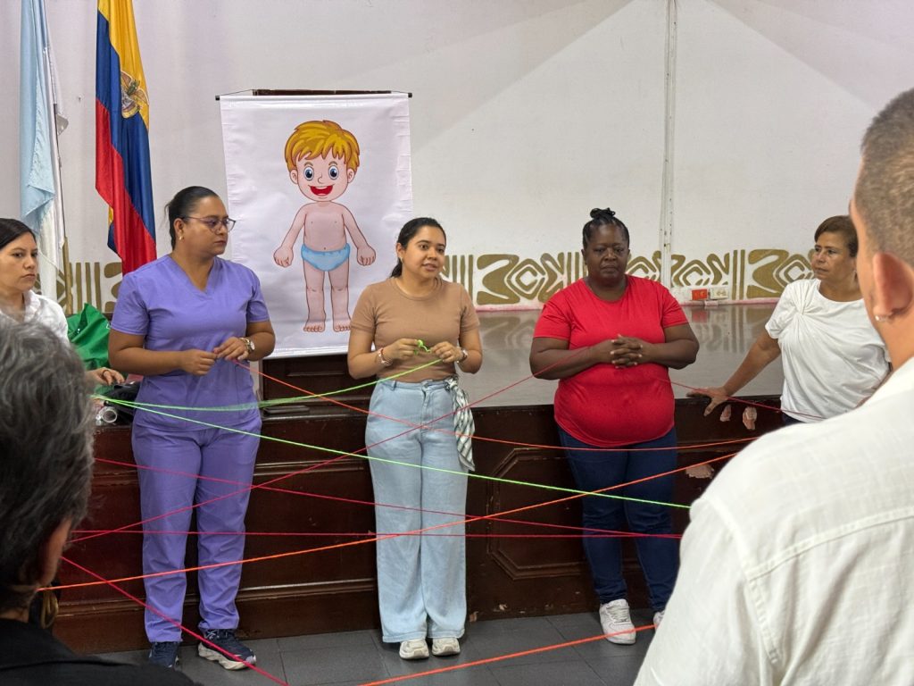 Fotografía de un grupo diverso de personas adultas participando en una dinámica grupal con hilos de colores cruzados, frente a un cartel con un dibujo infantil del cuerpo humano y una bandera de Colombia al fondo.