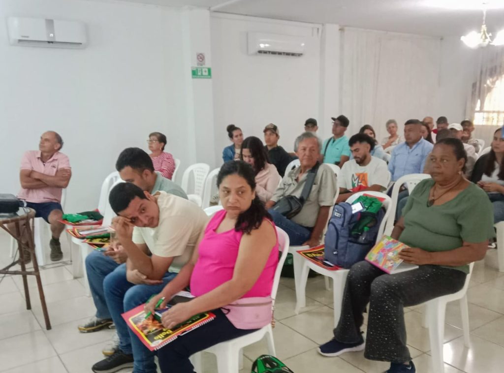 Fotografía de un grupo diverso de personas adultas sentadas en sillas plásticas blancas dentro de un salón, prestando atención durante una sesión informativa o taller comunitario.