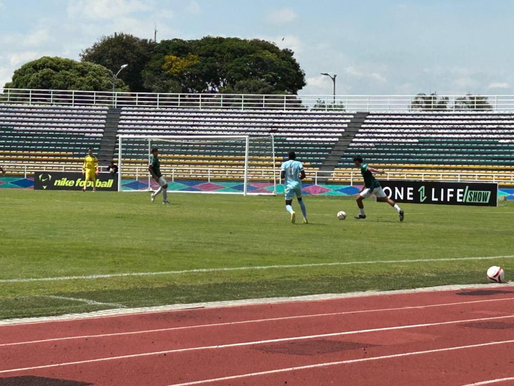 Personas jóvenes disputando un partido de fútbol en la cancha del estadio de Palmira, con la pista atlética en primer plano y las tribunas de fondo en un día soleado