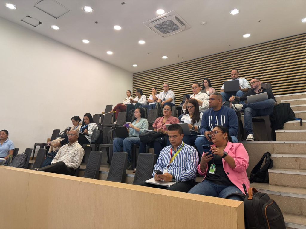 Asistentes a una jornada de capacitación o reunión institucional sentados en las graderías de un auditorio, prestando atención y utilizando sus computadores y dispositivos móviles.
