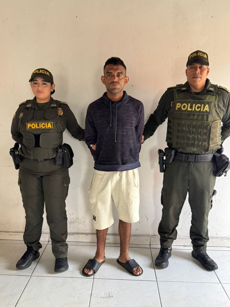 dos integrantes de la Policía Nacional de Colombia —una mujer a la izquierda y un hombre a la derecha— custodiando a una persona detenida en el centro. Las tres personas se encuentran de pie frente a una pared blanca y lisa.