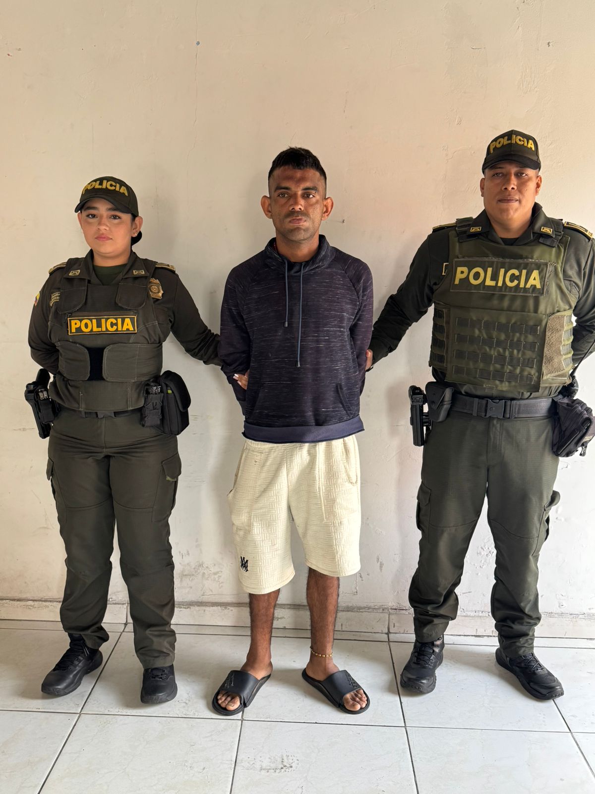 dos integrantes de la Policía Nacional de Colombia —una mujer a la izquierda y un hombre a la derecha— custodiando a una persona detenida en el centro. Las tres personas se encuentran de pie frente a una pared blanca y lisa.