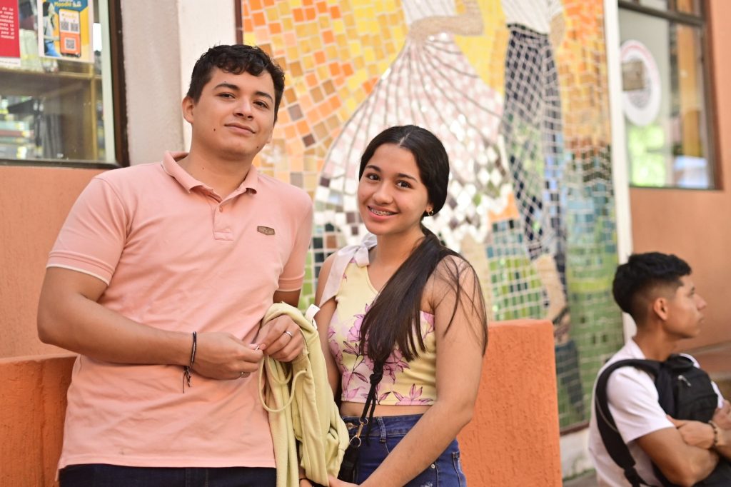 Dos personas jóvenes sonríen a la cámara durante una jornada institucional en Palmira, con un mural artístico de mosaicos al fondo y otra persona sentada de perfil.