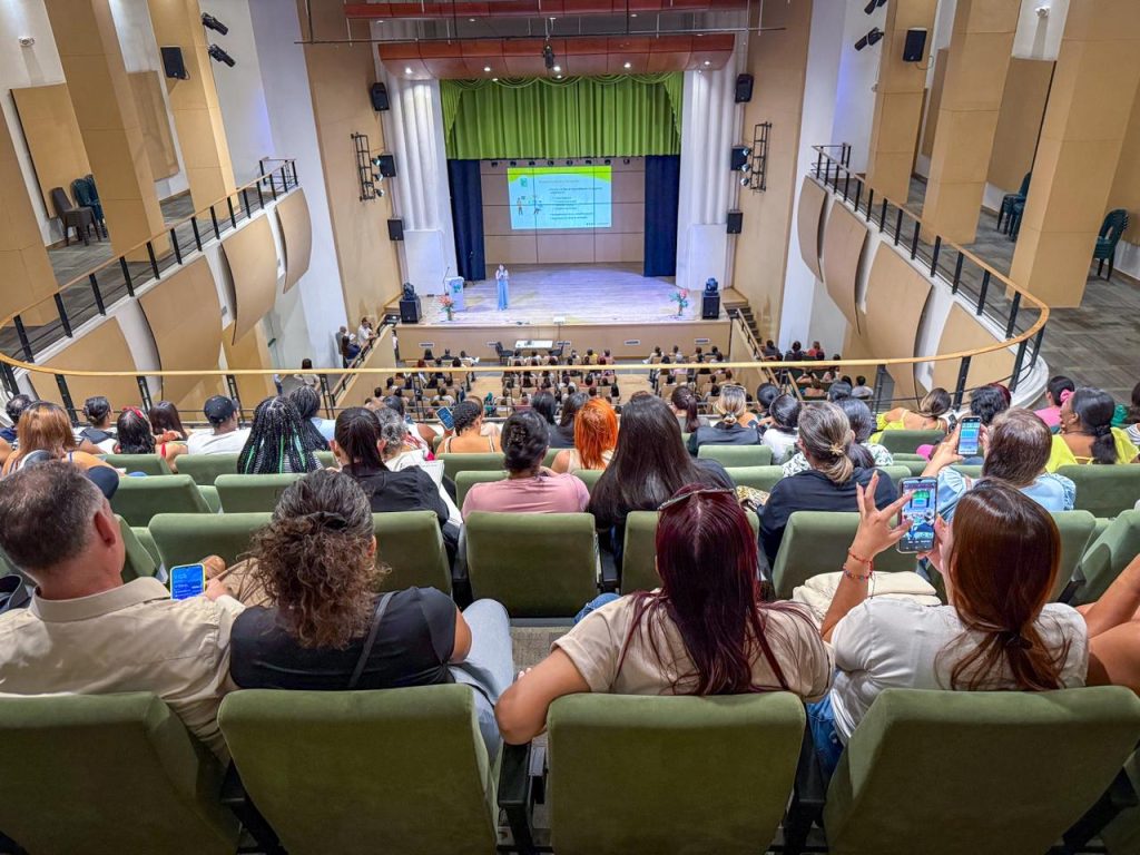 Vista aérea en picado desde los asientos superiores de un auditorio moderno. Un público diverso ocupa las butacas de color verde, prestando atención a una persona expositora que habla en el escenario frente a una pantalla de proyección. Algunas personas del público sostienen sus teléfonos móviles.
